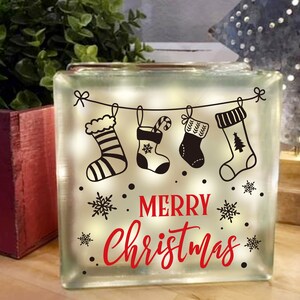 May include: A clear glass block with a white background and a black and white design of Christmas stockings hanging from a string with the text "Merry Christmas" in red letters.