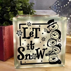 Op de afbeelding: Een witte glazen blok met een zwart silhouet van sneeuwpoppen en de tekst "Let it Snow".