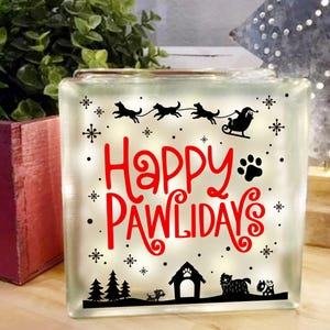 May include: A white light-up block with the text "Happy Pawlidays" in red lettering. The block features a silhouette of a Santa Claus riding in a sleigh pulled by dogs. There are also silhouettes of dogs and snowflakes on the block.