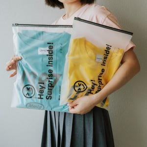 May include: Two clear plastic zippered bags with the text "Hey! Surprise inside!" printed on them. One bag contains a light blue t-shirt and the other contains a yellow t-shirt.