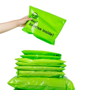 Könnte beinhalten: Ein Stapel leuchtend grüner Poly-Versandtaschen mit dem Text "Hey! Surprise inside!" auf der obersten Tasche.
