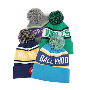 Puede incluir: Cuatro gorros de punto coloridos con pompones. Los gorros son en tonos de azul, verde, gris y amarillo, cada uno con una palabra o nombre diferente en letras blancas. Los gorros tienen un puño acanalado y un pompón en la parte superior.