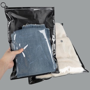 May include: Two clear, black-trimmed storage bags. One bag contains blue denim jeans, and the other contains a white, fluffy garment. The bags are sealed with a black zipper and a metal ring.