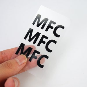 K&ouml;nnte beinhalten: Drei transparente Bl&auml;tter mit den Buchstaben "MFC" in Schwarz auf jedem Blatt.