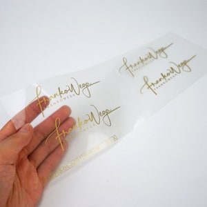 K&ouml;nnte beinhalten: Transparente Folie mit zwei goldenen Folienaufklebern mit dem Text "FrankoWega" in einer handschriftlichen Schriftart. Der Text "FrankoWega" wird zweimal auf der Folie wiederholt. Die Folie enth&auml;lt auch den Text "#2018-01-A6911[ ]限 101-X".