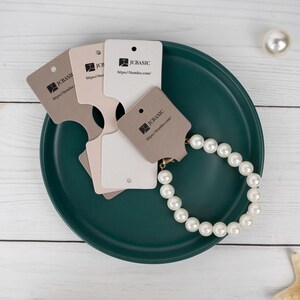 May include: A pearl bracelet and several product tags are arranged on a dark teal plate. The tags are in shades of beige and white, with the "JCBASIC" logo and website address printed on them. A single pearl and a starfish are also visible.