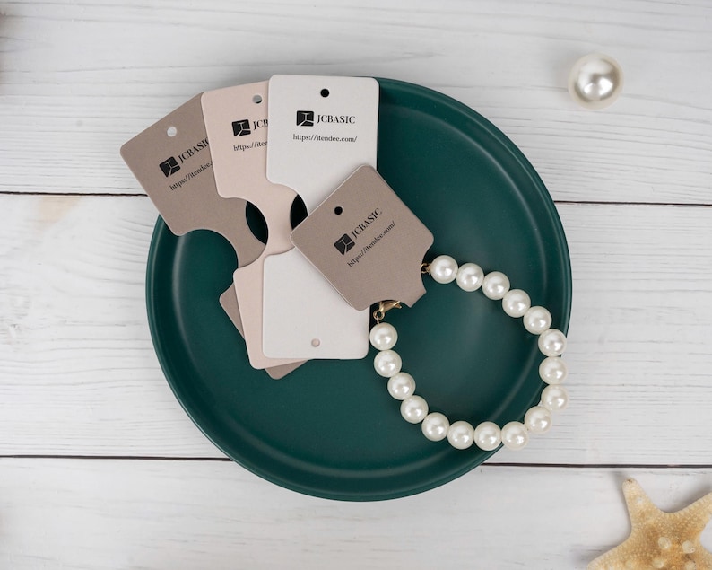 May include: A pearl bracelet and several product tags are arranged on a dark teal plate. The tags are in shades of beige and white, with the "JCBASIC" logo and website address printed on them. A single pearl and a starfish are also visible.