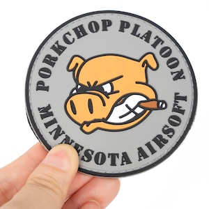 Könnte beinhalten: Ein runder Aufnäher mit dem Text "PORKCHOP PLATOON MINNESOTA AIRSOFT" in schwarzer Schrift auf grauem Hintergrund. Der Aufnäher zeigt ein Cartoon-Schwein mit einer Zigarre im Mund, mit einem wütenden Gesichtsausdruck.