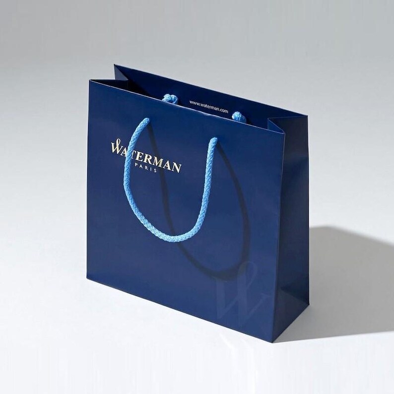 May include: A blue shopping bag with a white logo that says "Waterman Paris" and a blue rope handle. The website address "www.waterman.com" is printed on the bag.