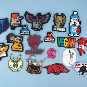 May include: A collection of colorful rubber patches with various designs. Includes a blue bear head, a black eagle with "BLCK PHNX" text, a dog with a baseball bat, and a "VEGAN" patch. Other patches feature military and sports themes.