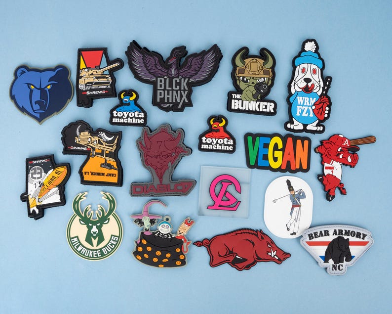 May include: A collection of colorful rubber patches with various designs. Includes a blue bear head, a black eagle with "BLCK PHNX" text, a dog with a baseball bat, and a "VEGAN" patch. Other patches feature military and sports themes.