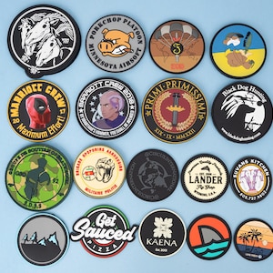 May include: A collection of round patches with various designs. Some feature cartoon characters, military emblems, and text like "Maximum Effort" and "Get Sauced Pizza." The patches are in various colors, including black, white, red, and blue.