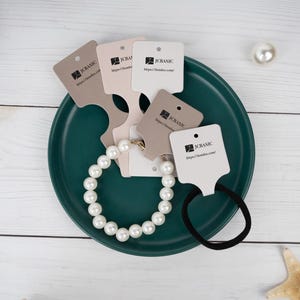 May include: A pearl bracelet and black hair tie rest on a teal plate, surrounded by several product tags. The tags are in various neutral colors and feature the text "JC BASIC" and a website address. A single pearl and starfish are also visible.
