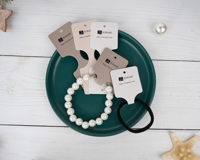 May include: A pearl bracelet and black hair tie rest on a teal plate, surrounded by several product tags. The tags are in various neutral colors and feature the text "JC BASIC" and a website address. A single pearl and starfish are also visible.