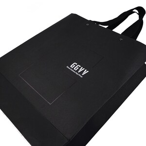 以下が含まれることがあります： 黒いショッピングバッグ。黒いハンドルと、白で「GGVV Glamour Gentlemen Vipe Vintage」の文字。バッグは長方形で、テクスチャ加工された素材で作られています。