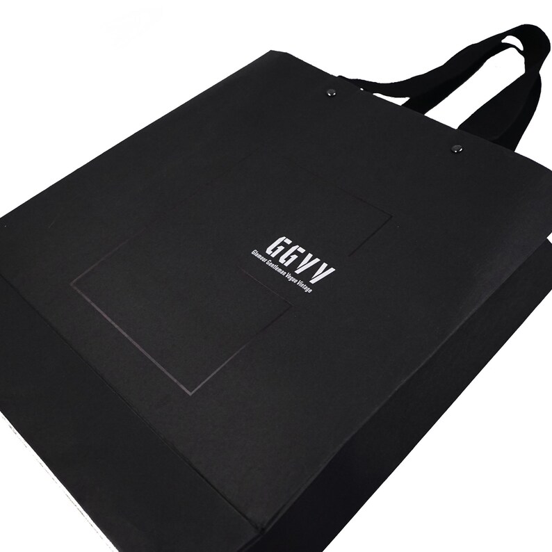 以下が含まれることがあります： 黒いショッピングバッグ。黒いハンドルと、白で「GGVV Glamour Gentlemen Vipe Vintage」の文字。バッグは長方形で、テクスチャ加工された素材で作られています。