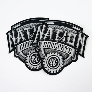 Könnte beinhalten: Zwei schwarze und silberne gestickte Patches mit dem Text "Nation Concrete" in einer stilisierten Schriftart. Die Patches haben ein kreisförmiges Design mit den Buchstaben "NC" in der Mitte.