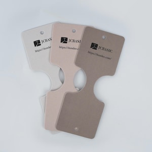 May include: Three necklace display cards in shades of beige and brown. Each card has a unique shape with a hole at the top and a cutout at the bottom. The cards feature the text "JCBasic" and a website address.