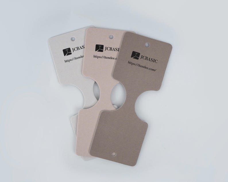 May include: Three necklace display cards in shades of beige and brown. Each card has a unique shape with a hole at the top and a cutout at the bottom. The cards feature the text "JCBasic" and a website address.