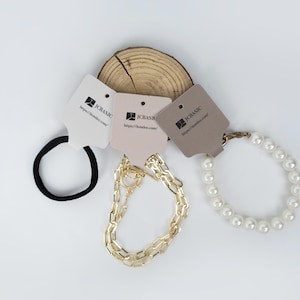 May include: An assortment of jewelry and hair accessories displayed on a white surface. The items include a black hair tie, a gold chain necklace, and a pearl bracelet. Each item has a tag with the text "JC BASIC" and a website address.