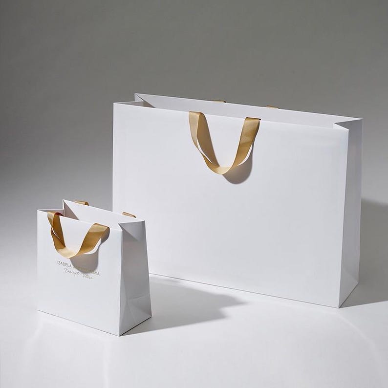 May include: Two white paper shopping bags with gold ribbon handles. The smaller bag has the text "GABBANA Concept Store" printed on the front.