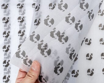 May include: White tissue paper with a repeating pattern of black globe shapes. The tissue paper is folded in half and held by a hand.