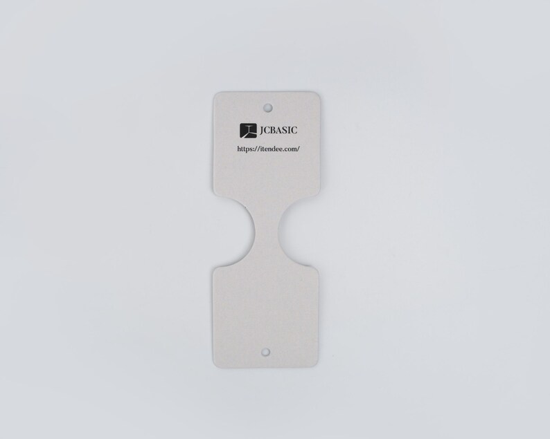 May include: A light gray, double-ended tag with a unique cutout design. The tag features the JCBASIC logo and website address. It has two small holes for attachment.