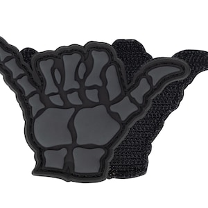 Könnte beinhalten: Ein schwarzer Aufnäher mit einer stilisierten Skeletthand im Shaka-Zeichen. Der Aufnäher hat ein strukturiertes, gummiartiges Aussehen mit einem Klettverschluss auf der Rückseite zur Befestigung. Das Design ist dunkelgrau.