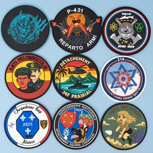 May include: A collection of nine round patches with various designs and text. The patches feature cartoon characters, military themes, and text such as "P-431", "REPARTO ARMI", and "HERE WE GOO". The patches are on a light blue background.