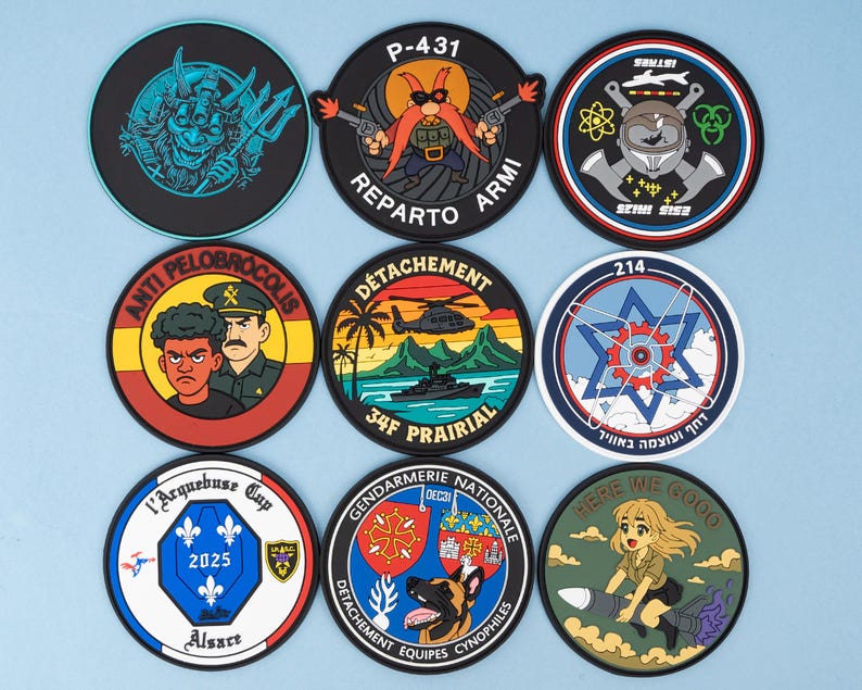 May include: A collection of nine round patches with various designs and text. The patches feature cartoon characters, military themes, and text such as "P-431", "REPARTO ARMI", and "HERE WE GOO". The patches are on a light blue background.