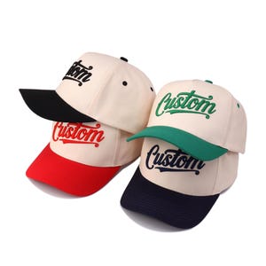 May include: Cream-coloured baseball caps with curved peaks. Each cap has the word "Custom" embroidered in a contrasting colour. One has a black peak, another a red peak, and the third has a green peak. The caps are stacked.