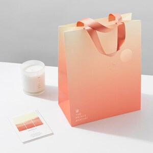 May include: A peach-colored shopping bag with a pink ribbon handle. The bag has a gradient design with a lighter peach color at the top and a darker peach color at the bottom. The bag has a white circle with a sun inside and a few white birds flying above it. The bag has the text "THE PRINCE AKATOKI" printed on the bottom.