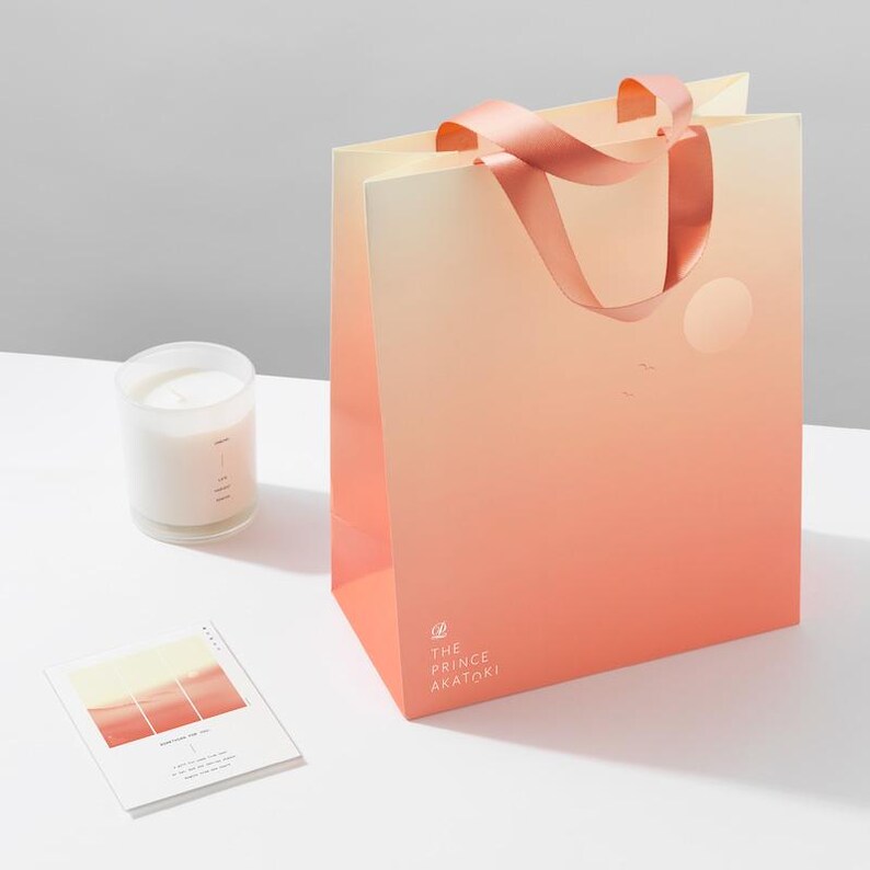 May include: A peach-colored shopping bag with a pink ribbon handle. The bag has a gradient design with a lighter peach color at the top and a darker peach color at the bottom. The bag has a white circle with a sun inside and a few white birds flying above it. The bag has the text "THE PRINCE AKATOKI" printed on the bottom.