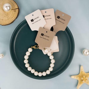May include: A pearl bracelet with a gold clasp rests on a teal plate, surrounded by several tags with the text "JCBASIC" and a website address. Additional pearls and a starfish are scattered around the plate, set against a light blue background.