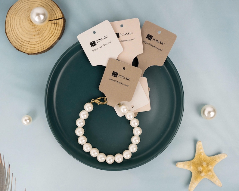 May include: A pearl bracelet with a gold clasp rests on a teal plate, surrounded by several tags with the text "JCBASIC" and a website address. Additional pearls and a starfish are scattered around the plate, set against a light blue background.