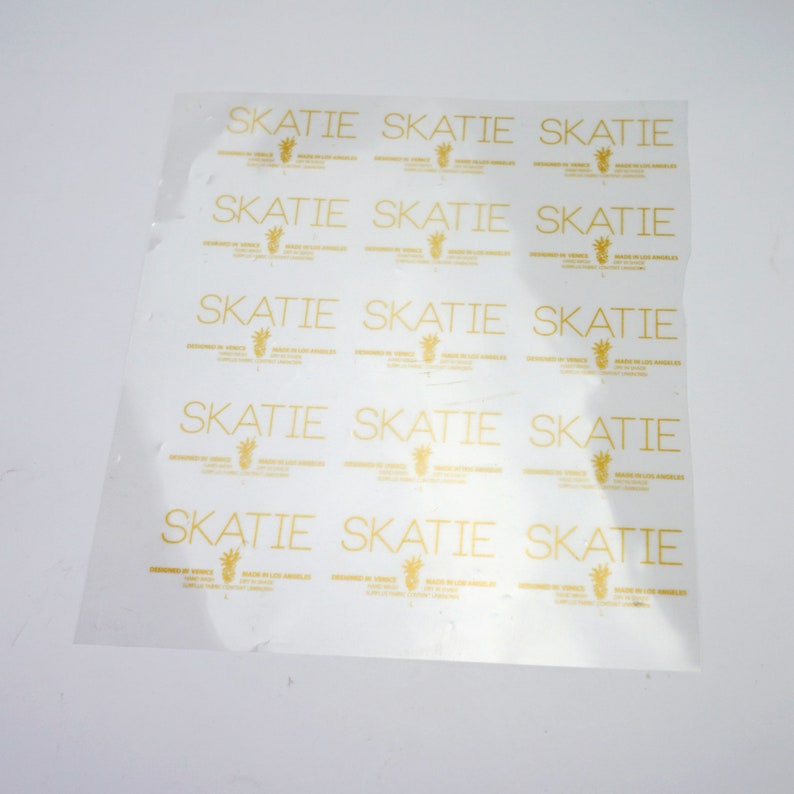 K&ouml;nnte beinhalten: Ein Blatt aus durchsichtigem Plastik mit dem Wort "SKATIE" 15 Mal in goldener Schrift wiederholt. Der Text ist in einer einfachen, serifenlosen Schriftart.