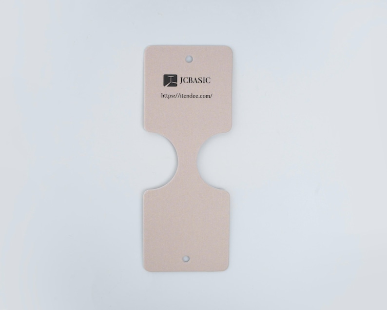 May include: A light beige product tag with a unique shape, featuring a logo and the text "JCBASIC" and "https://itendee.com/". The tag has a hole at the top and bottom for attachment. The tag is made of a sturdy material.