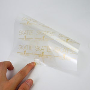 K&ouml;nnte beinhalten: Ein Blatt aus klarem, transparentem, selbstklebendem Papier mit einem sich wiederholenden Muster des Wortes "SKATTIE" in goldener Schrift. Die Texte "DESIGNED IN NYC" und "MADE IN LOS ANGELES" sind ebenfalls in goldener Schrift gedruckt.