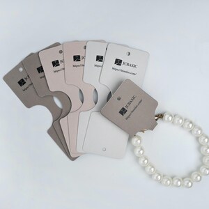 May include: A pearl bracelet displayed with several neutral-toned jewelry display cards. The cards have a unique shape with a cutout and a small hole for hanging. Each card features the text "JCBASIC" and a website address.