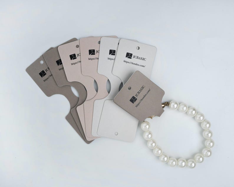 May include: A pearl bracelet displayed with several neutral-toned jewelry display cards. The cards have a unique shape with a cutout and a small hole for hanging. Each card features the text "JCBASIC" and a website address.