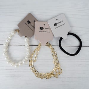 May include: An assortment of jewelry and hair accessories on a white wooden surface. The items include a pearl bracelet, a gold chain bracelet, and a black hair tie. Each item has a tag with the text "JCBASIC" and a website address.