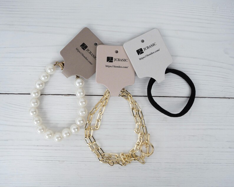 May include: An assortment of jewelry and hair accessories on a white wooden surface. The items include a pearl bracelet, a gold chain bracelet, and a black hair tie. Each item has a tag with the text "JCBASIC" and a website address.