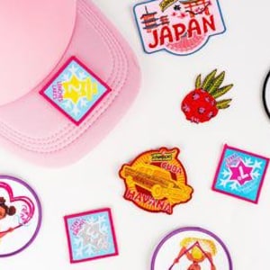 May include: A pink baseball cap with several embroidered patches scattered around it. The patches feature various designs, including a pineapple, a car, and the word "Japan". The patches are in various colors, including red, blue, and yellow.