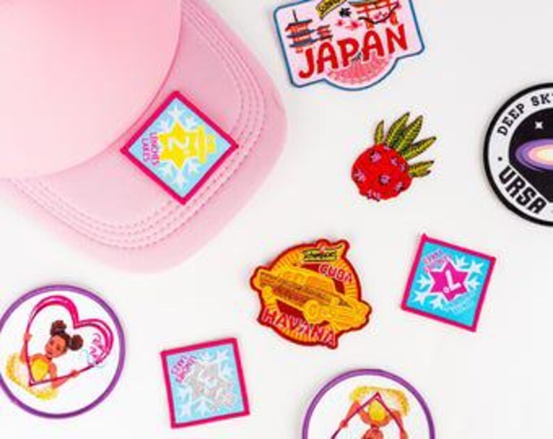 May include: A pink baseball cap with several embroidered patches scattered around it. The patches feature various designs, including a pineapple, a car, and the word "Japan". The patches are in various colors, including red, blue, and yellow.