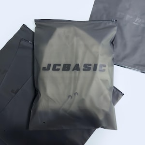 Puede incluir: Se muestran varias bolsas de plástico grises translúcidas con cremalleras. Las bolsas tienen el texto "JC BASIC" impreso en color gris oscuro. Las bolsas parecen estar vacías y están sobre una superficie blanca.