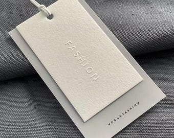 May include: White rectangular tag with the word "FASHION" embossed in the center. The tag is attached to a gray fabric with the text "VOGUEFASHION" printed on the bottom right corner.