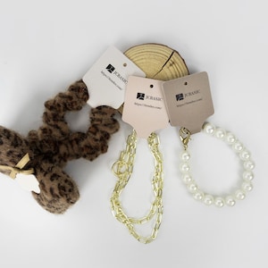 May include: An assortment of accessories, including a leopard print scrunchie with a bow, a gold chain necklace, and a pearl bracelet. Each item has a tag with the text "JCBASIC" and a website address. The items are arranged on a white surface.