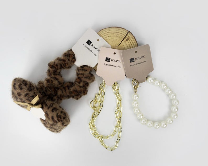 May include: An assortment of accessories, including a leopard print scrunchie with a bow, a gold chain necklace, and a pearl bracelet. Each item has a tag with the text "JCBASIC" and a website address. The items are arranged on a white surface.