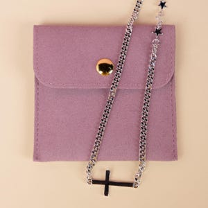 Puede incluir: Un collar plateado con un colgante de cruz y detalles de estrellas descansa sobre una bolsa malva con cierre de botón dorado. El collar presenta un diseño de cadena de eslabones.