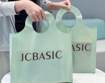 Könnte beinhalten: Zwei hellgrüne Einkaufstaschen mit kreisförmigen Griffen. Die Taschen tragen den braunen Aufdruck "JC BASIC". Die Taschen werden von einer Person gehalten, und der Hintergrund ist neutral gefärbt.
